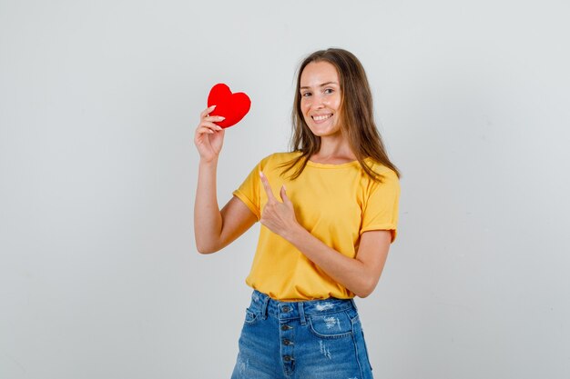 Dedo acusador femenino joven en corazón rojo en camiseta, pantalones cortos y mirando alegre