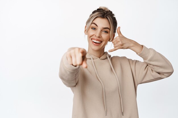 Dedo acusador joven mujer rubia, mostrando gesto de teléfono móvil, me llamo señal, sonriendo feliz, de pie en blanco.