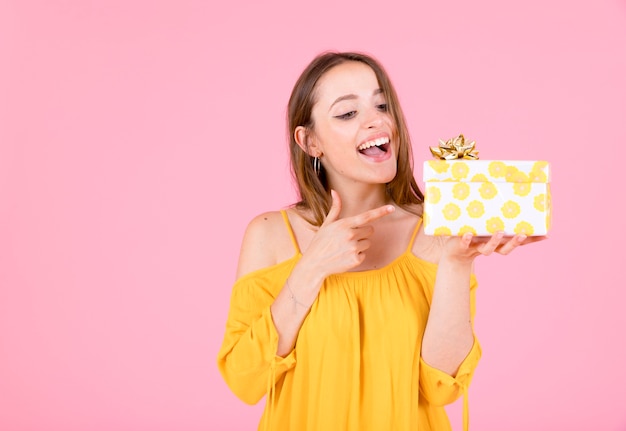 Dedo acusador de mujer joven feliz a la caja de regalo sobre fondo rosa