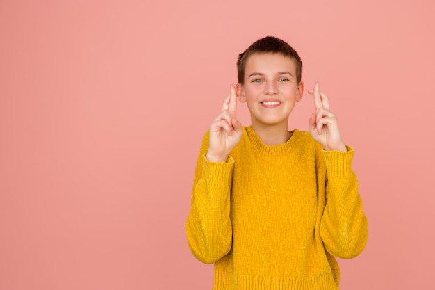 Dedos cruzados. Retrato de niña caucásica sobre fondo de estudio rosa coral con espacio de copia para anuncio. Hermosa modelo en suéter. Concepto de emociones humanas, expresión facial, ventas, publicidad, moda.