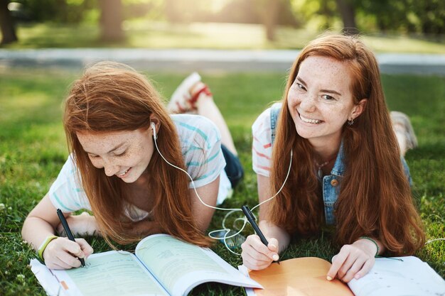 Deja de jugar y haz la tarea Dos hermanas pelirrojas relajadas y despreocupadas con pecas tiradas en el césped en el parque escribiendo notas para la universidad compartiendo auriculares escuchando canciones juntas riendo