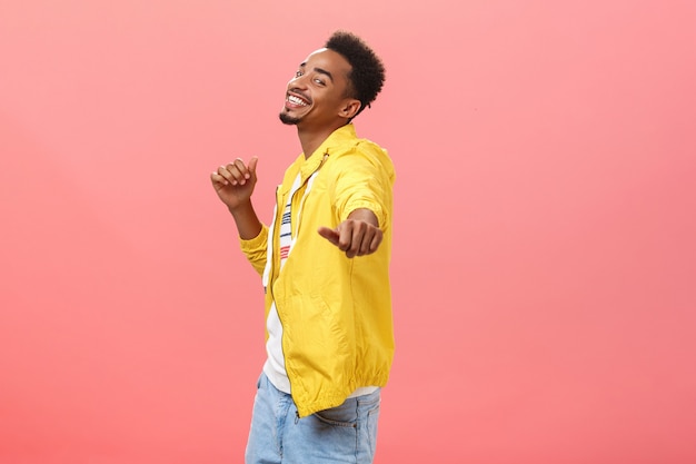 Dejarse llevar por la alegría y las emociones positivas. Retrato de alegre elegante feliz chico de piel oscura con corte de pelo afro bailando apuntando a la cámara con la mano y sonriendo felizmente