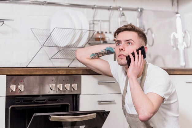 Delantal que lleva masculino joven serio que habla en el teléfono móvil que mira lejos