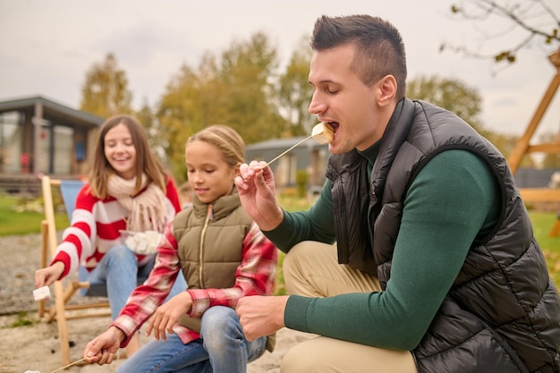 Delicadeza, placer. Perfil de un hombre de mediana edad con los ojos cerrados mordiendo con placer malvaviscos en una chica de palo y una mujer detrás de una fogata en el patio de la casa el día de otoño