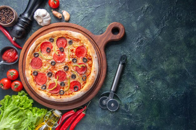 Deliciosa pizza casera en la tabla de cortar de madera, tomates, salsa de tomate, ajo, paquete verde, botella de aceite en el lado derecho sobre una superficie oscura