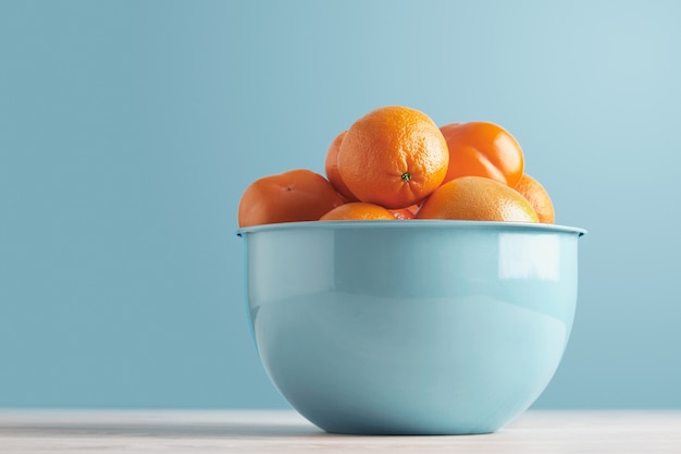 Deliciosas frutas frescas maduras y cítricos en un recipiente azul metálico aislado en una mesa blanca sobre fondo azul pastel: caqui, ciruela, mandarina, naranja, pomelo, pomelo