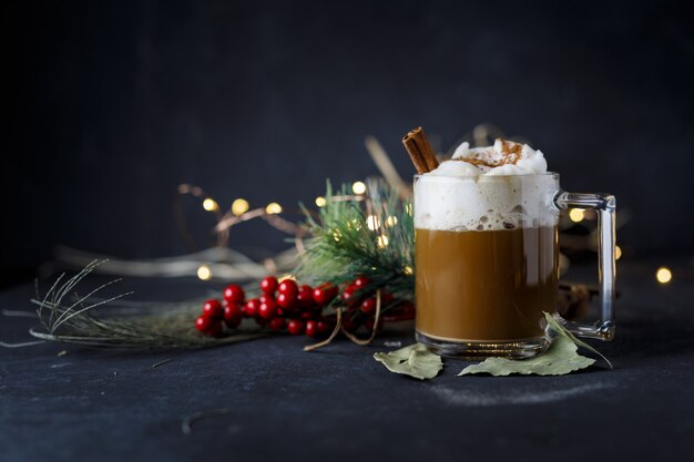 Delicioso café navideño con canela y espuma, junto a acebos sobre una superficie oscura