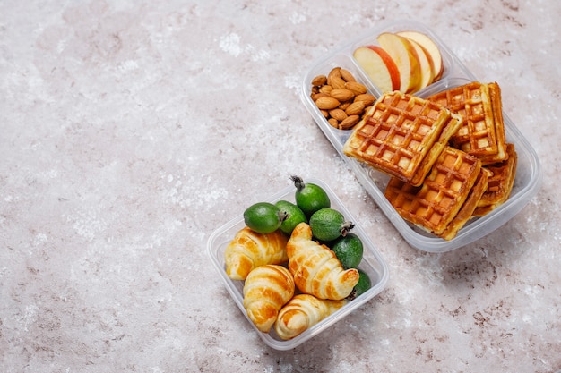 Delicioso desayuno con almendras, rodajas de manzana roja, gofres, cruasanes en una lonchera de plástico a la luz