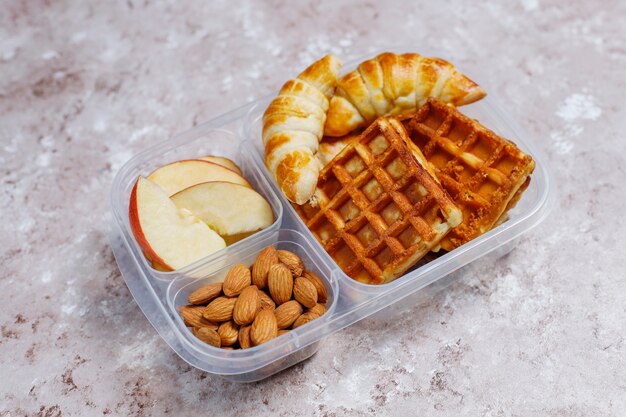 Delicioso desayuno con almendras, rodajas de manzana roja, gofres, cruasanes en una lonchera de plástico a la luz