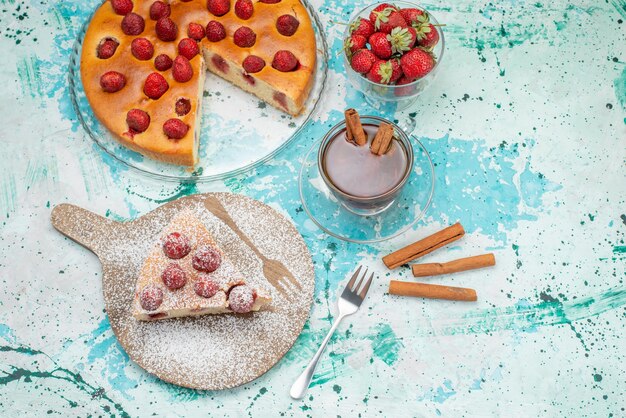 Delicioso pastel de fresa en rodajas y todo un delicioso pastel de azúcar en polvo con té en un escritorio azul brillante, masa para hornear dulce de baya