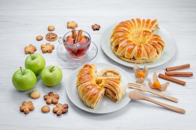 Deliciosos pasteles en rodajas dentro de la placa con relleno junto con té de manzanas verdes y galletas en el escritorio blanco, pastelería cookie biscuit dulce