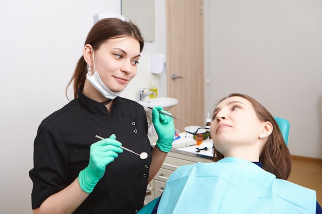 Dentista con experiencia y atractiva mujer morena joven sosteniendo herramientas dentales mientras revisa los dientes de la paciente para caries