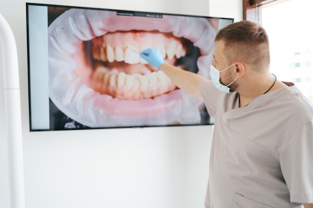 Dentista en máscara médica apuntando a los dientes del paciente en una pantalla grande que explica las fases del tratamiento