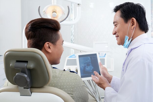 Dentista masculino asiático que muestra la imagen de rayos X de la mandíbula en la tableta al paciente