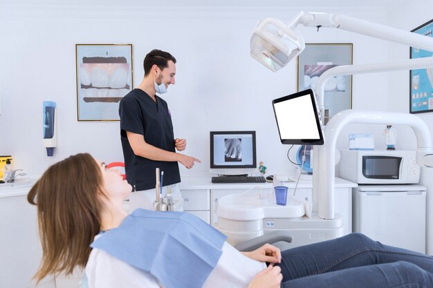Un dentista masculino que explica una radiografía de los dientes en la pantalla al paciente femenino que miente en la silla del dentista