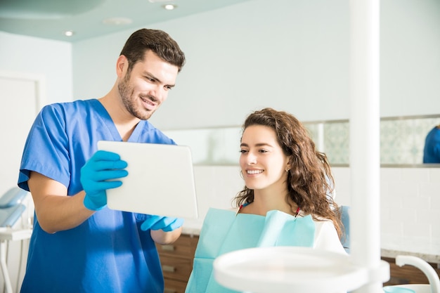 Dentista de mediana edad que muestra una tableta digital a una paciente durante el tratamiento en una clínica dental
