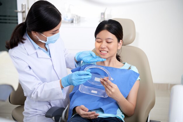 Dentista mujer asiática dando espejo de mano al paciente y demostrando el resultado del tratamiento