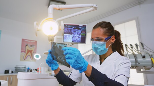 Dentista personalizando la luz sobre el sillón y analizando la radiografía de los dientes antes de la cirugía. Médico de odontología encendiendo la lámpara y examinando a la persona con máscara de protección y gafas, enfermera ayudándola