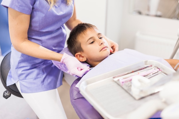 Dentista que se sienta cerca del muchacho que se inclina en silla dental en clínica