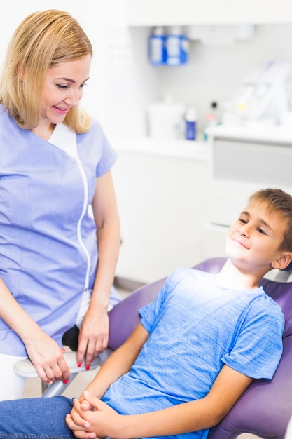 Dentista de sexo femenino feliz que mira al muchacho que se sienta en silla dental