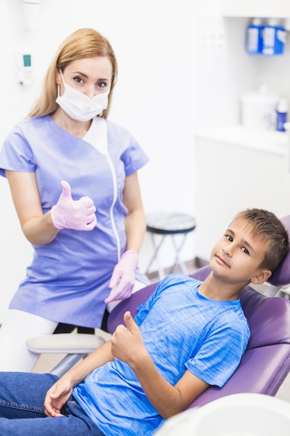 Dentista de sexo femenino joven y muchacho que gesticulan los pulgares para arriba en clínica