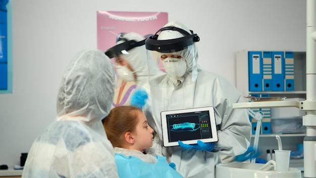 Dentista con traje de ppe apuntando en la pantalla digital que explica la radiografía a la madre de la paciente. Equipo médico y pacientes que usan overoles de protección facial, mascarillas, guantes, mostrando radiografía usando un cuaderno