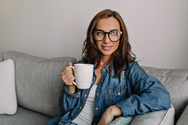 Dentro de retrato de joven atractiva mujer elegante con piel bronceada y cabello castaño con anteojos mirando una ventana