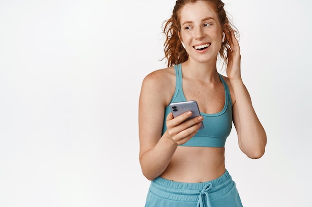 Deporte y fitness Sonriente atleta femenina escuchando música en auriculares con teléfono móvil de pie en ropa deportiva contra el fondo blanco.