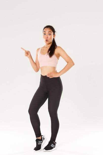 Deporte, gimnasio y concepto de cuerpo sano. Longitud total de chica fitness asiática morena linda sorprendida en ropa deportiva, mirando sorprendido, señalando con el dedo hacia la izquierda, mostrando precios increíbles para equipos de gimnasio.