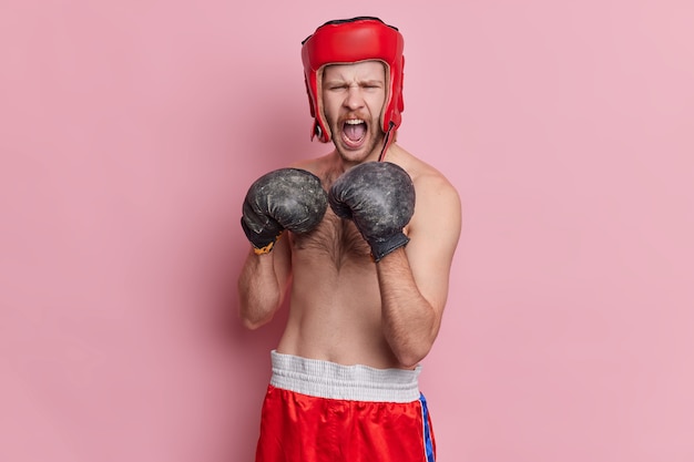 Deporte de personas y concepto de estilo de vida saludable. Boxeador masculino emocional grita airadamente lleva casco protector, guantes de boxeo y pantalones cortos listos para la pelea