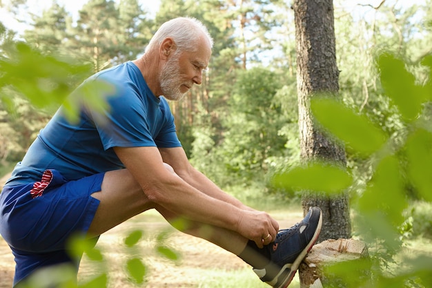 Deportes, determinación, resistencia y actividad. Vista lateral del hombre mayor barbudo musculoso con estilo posando en la naturaleza salvaje, atando cordones en zapatillas de deporte, listo para correr. Enfoque selectivo en el hombre