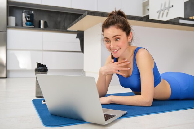 Foto gratuita deportista acostada en la alfombra de fitness y mirando sorprendida en la pantalla de la computadora portátil hablando con alguien en