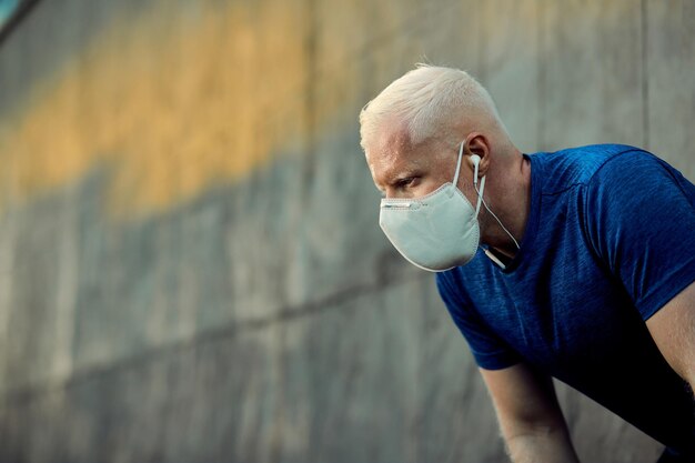 Deportista albino sudoroso recuperando el aliento después de correr con mascarilla protectora al aire libre