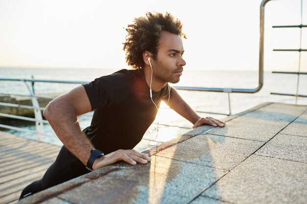 Deportista confiado con peinado tupido haciendo ejercicios en el muelle temprano en la mañana. Colocando los brazos en la plataforma mientras escucha música.