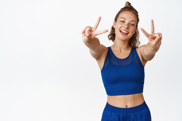 Deportista feliz y saludable muestra la paz v-sign y sonriendo, vistiendo ropa deportiva. Corredor de fitness femenino saludando, de pie sobre blanco