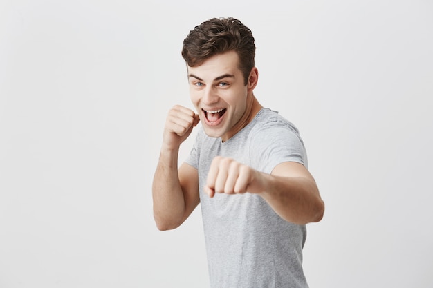 Deportista joven musculoso seguro y positivo con cabello oscuro y ojos azules vestido con camisa gris sonriendo ampliamente, sosteniendo los puños al frente, listo para nuevos desafíos, demostrando su fuerza