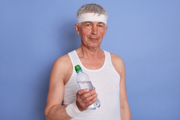 Deportista masculino senior de pelo blanco, teniendo un descanso entre series durante el entrenamiento, sosteniendo una botella de agua, posando con ropa blanca aislada.