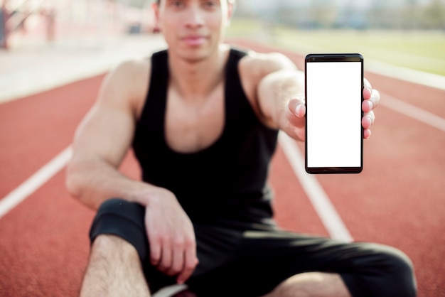 Deportista masculino sentado en la pista de carreras mostrando la pantalla del teléfono móvil