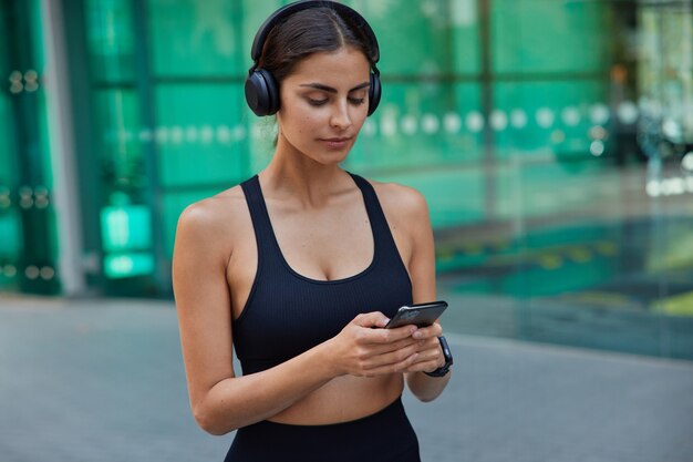 Deportista morena seria vestida con mensajes de texto de tipo superior recortado escucha la pista de audio en los auriculares tiene entrenamiento físico borroso