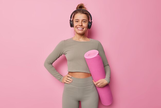 Deportista motivada con cabello peinado en ropa deportiva que lleva karemat enrollado se prepara para el entrenamiento físico escucha música a través de auriculares inalámbricos disfruta de su lista de reproducción favorita aislada sobre fondo rosa.