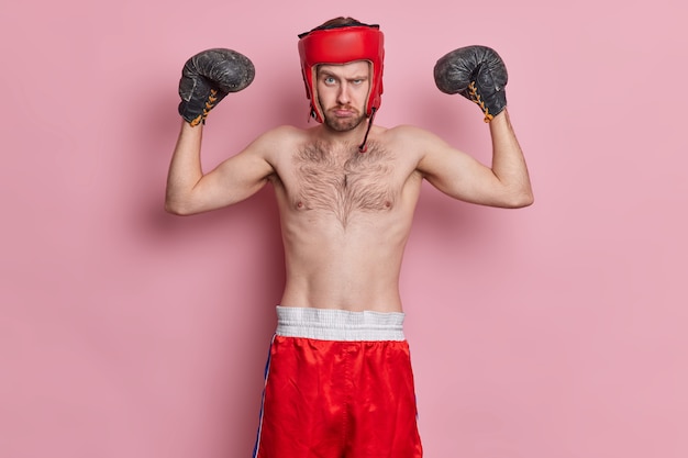 Foto gratuita deportista motivado disfruta del boxeo usa gorro de protección, guantes, levanta los brazos, muestra músculos, tiene un cuerpo delgado y el torso desnudo parece serio