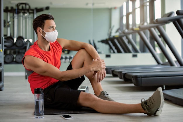 Deportista pensativo con mascarilla protectora relajándose en el suelo en el club de salud