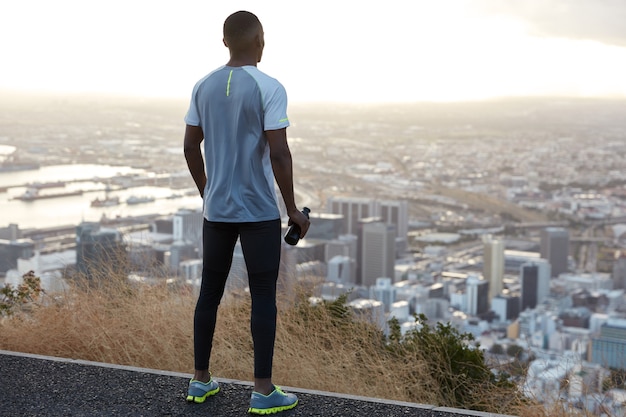 Deportista de piel oscura con ropa deportiva, se aparta, bebe agua de la botella, usa zapatillas, se para en lo alto y disfruta de la vista panorámica de la ciudad con rascacielos desde arriba, hace ejercicio al aire libre en el campo