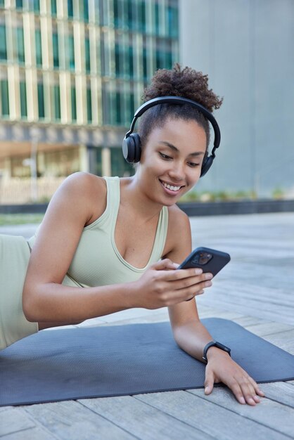 Deportista positiva escucha música a través de auriculares navega por Internet en el teléfono móvil vestida con chándal se apoya en la alfombra de fitness va a hacer deporte contra el fondo borroso de la ciudad Deporte y tecnología