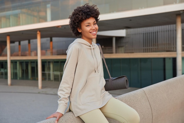 La deportista positiva usa sudadera lleva colchoneta de fitness tiene buen humor después de practicar yoga o hacer ejercicio se sienta relajada al aire libre en un entorno urbano tiene expresión feliz gente y estilo de vida activo