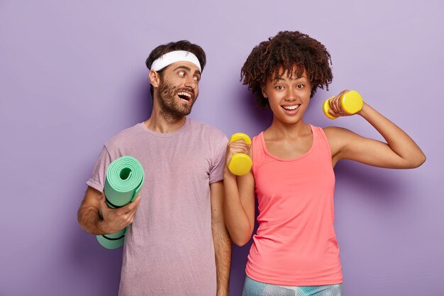 Deportista positivo usa diadema y camiseta, sostiene una alfombra de fitness arrugada, mira con alegría a la novia que levanta los brazos con pesas, hacen ejercicio juntos