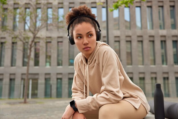 Deportista reflexiva descansa después de que el entrenamiento de cardio duro se sienta al aire libre y se concentra vestida con una sudadera con capucha escucha música en auriculares