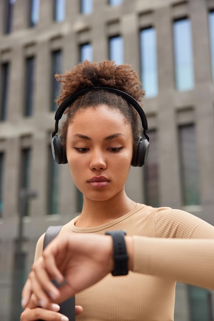 La deportista rizada seria comprueba los resultados del entrenamiento en el reloj inteligente escucha música en auriculares inalámbricos vestida con ropa deportiva