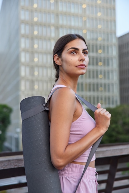 Deportista en ropa deportiva lleva colchoneta enrollada en el hombro va a tener ejercicios al aire libre concentrados en poses de raspadores de la ciudad