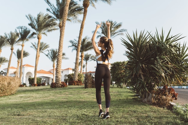 Deportista sexy estirando las manos sobre la hierba en la ciudad tropical. Mañana soleada, estado de ánimo alegre, motivación, entrenamiento, escalofriante con los ojos cerrados, estilo de vida saludable, fitness, modelo atractivo